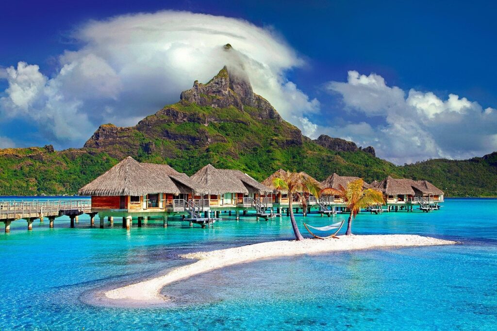 Überwasserbungalows mit Strohdächern vor einem Berg mit Wolken, türkisfarbenes Wasser und kleine Sandinsel mit Palmen und Hängematte