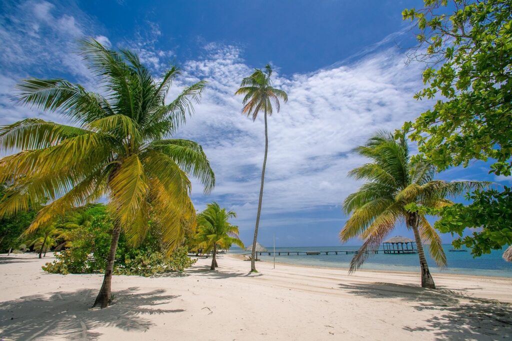 Strand mit weißem Sand, Palmen und blauem Himmel mit Wolken, im Hintergrund ein Steg ins Meer